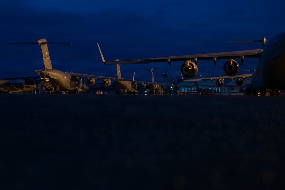 A low-angle nighttime photograph of multiple large military transport aircraft parked on a flight line or tarmac during twilight hours. The deep blue sky indicates either dusk or dawn conditions. At least two to three large four-engine military cargo aircraft are visible, consistent with C-17 Globemaster III or similar heavy-lift transports.