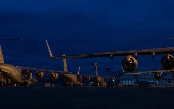 A low-angle nighttime photograph of multiple large military transport aircraft parked on a flight line or tarmac during twilight hours. The deep blue sky indicates either dusk or dawn conditions. At least two to three large four-engine military cargo aircraft are visible, consistent with C-17 Globemaster III or similar heavy-lift transports.