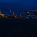 A low-angle nighttime photograph of multiple large military transport aircraft parked on a flight line or tarmac during twilight hours. The deep blue sky indicates either dusk or dawn conditions. At least two to three large four-engine military cargo aircraft are visible, consistent with C-17 Globemaster III or similar heavy-lift transports.