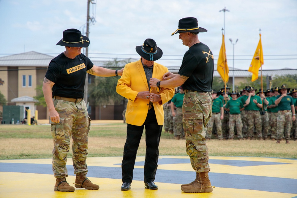 A military ceremony is taking place on a painted parade ground, where two soldiers in U.S. Army cavalry uniforms are assisting a civilian man in a bright yellow blazer, likely pinning an item or adjusting his attire. The setting appears to be a formal unit ceremony, possibly an induction, award, or honorary recognition event. A formation of soldiers in green and camouflage uniforms stands in the background alongside yellow cavalry guidon flags.