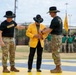 A military ceremony is taking place on a painted parade ground, where two soldiers in U.S. Army cavalry uniforms are assisting a civilian man in a bright yellow blazer, likely pinning an item or adjusting his attire. The setting appears to be a formal unit ceremony, possibly an induction, award, or honorary recognition event. A formation of soldiers in green and camouflage uniforms stands in the background alongside yellow cavalry guidon flags.