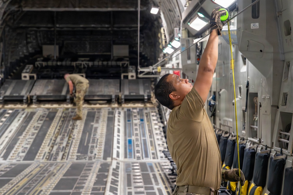 Two military personnel are working inside the cargo hold of a large military transport aircraft, likely a C-17 Globemaster III. The foreground figure is reaching upward with both hands to attach or adjust a strap or cable to an overhead anchor point, while a second figure works in the background near the rear ramp. The interior shows the characteristic roller-equipped cargo floor and tie-down rail systems typical of heavy military airlift aircraft.