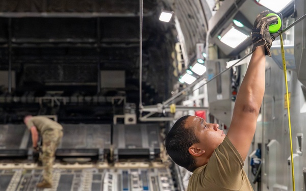 Two military personnel are working inside the cargo hold of a large military transport aircraft, likely a C-17 Globemaster III. The foreground figure is reaching upward with both hands to attach or adjust a strap or cable to an overhead anchor point, while a second figure works in the background near the rear ramp. The interior shows the characteristic roller-equipped cargo floor and tie-down rail systems typical of heavy military airlift aircraft.