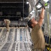 Two military personnel are working inside the cargo hold of a large military transport aircraft, likely a C-17 Globemaster III. The foreground figure is reaching upward with both hands to attach or adjust a strap or cable to an overhead anchor point, while a second figure works in the background near the rear ramp. The interior shows the characteristic roller-equipped cargo floor and tie-down rail systems typical of heavy military airlift aircraft.