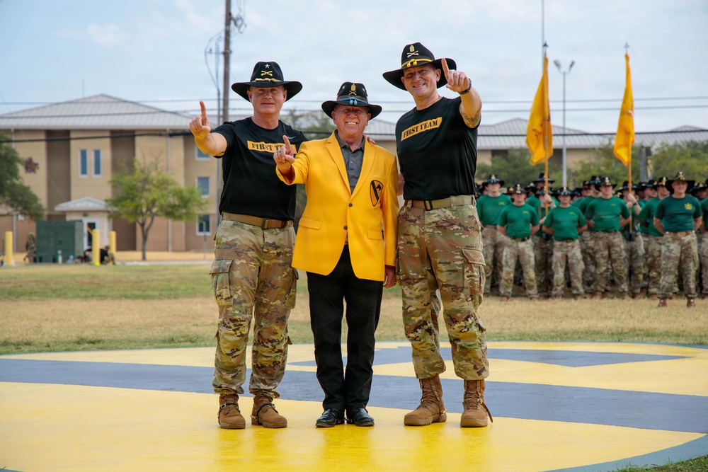 Three individuals pose for a formal photo on a painted yellow and black surface at what appears to be a military installation. Two soldiers in camouflage uniforms and cavalry Stetson hats flank a civilian wearing a distinctive gold blazer with a 1st Cavalry Division patch. All three are making a hand gesture (index finger raised) while formations of soldiers stand at attention in the background.