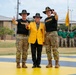 Three individuals pose for a formal photo on a painted yellow and black surface at what appears to be a military installation. Two soldiers in camouflage uniforms and cavalry Stetson hats flank a civilian wearing a distinctive gold blazer with a 1st Cavalry Division patch. All three are making a hand gesture (index finger raised) while formations of soldiers stand at attention in the background.