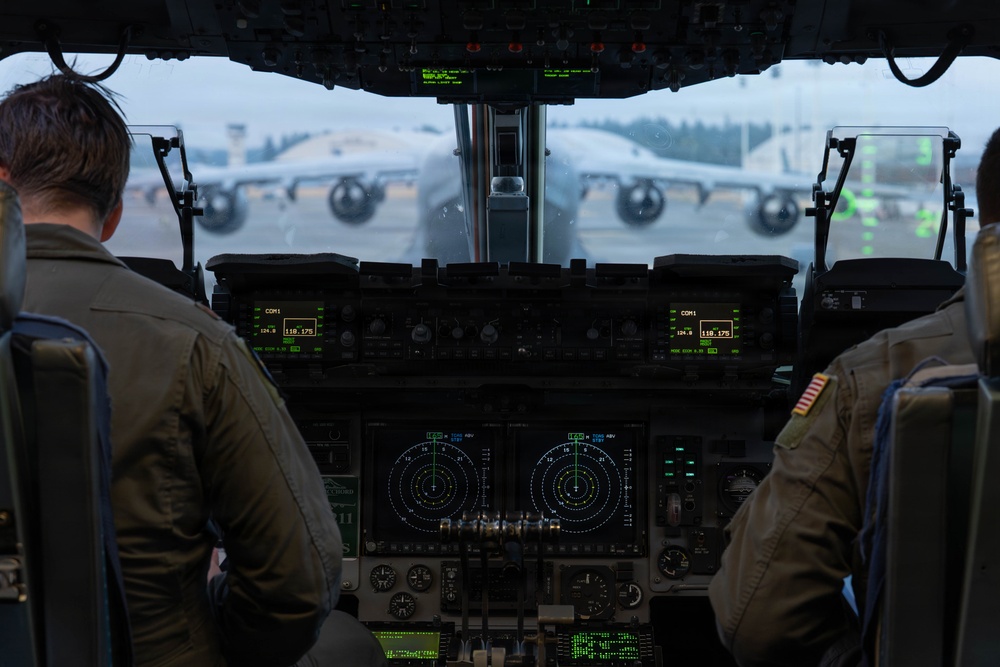 The image shows the interior cockpit view of a military transport aircraft, likely a C-17 Globemaster III, with two flight crew members seated at their stations. The cockpit features modern glass avionics displays including navigation/radar screens and communication panels. Through the windscreen, another large military transport aircraft is visible on the tarmac in overcast conditions.