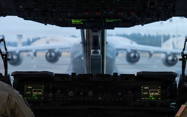 The image shows the interior cockpit view of a military transport aircraft, likely a C-17 Globemaster III, with two flight crew members seated at their stations. The cockpit features modern glass avionics displays including navigation/radar screens and communication panels. Through the windscreen, another large military transport aircraft is visible on the tarmac in overcast conditions.
