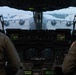 The image shows the interior cockpit view of a military transport aircraft, likely a C-17 Globemaster III, with two flight crew members seated at their stations. The cockpit features modern glass avionics displays including navigation/radar screens and communication panels. Through the windscreen, another large military transport aircraft is visible on the tarmac in overcast conditions.
