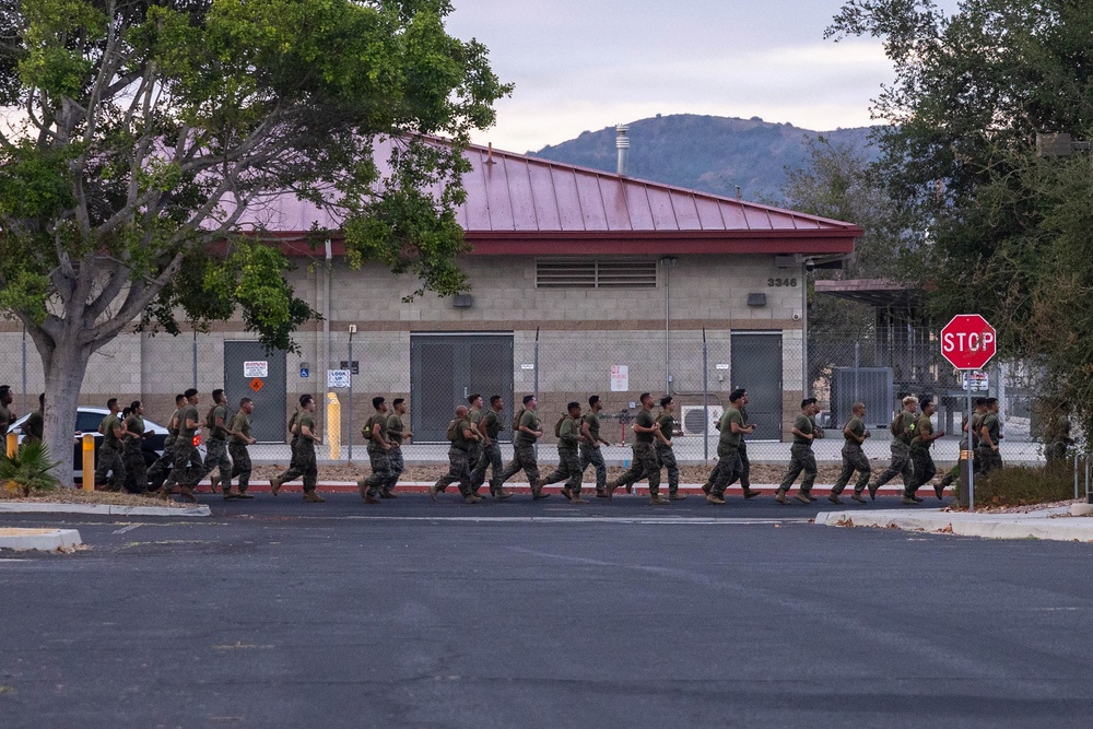 A formation of military personnel in camouflage uniforms are conducting a group run along a paved road on what appears to be a military installation. The building in the background bears the number 3346 and features a red metal roof with concrete block construction. The setting includes trees, hills in the background, and a stop sign, consistent with a U.S. military base environment.