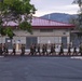 A formation of military personnel in camouflage uniforms are conducting a group run along a paved road on what appears to be a military installation. The building in the background bears the number 3346 and features a red metal roof with concrete block construction. The setting includes trees, hills in the background, and a stop sign, consistent with a U.S. military base environment.