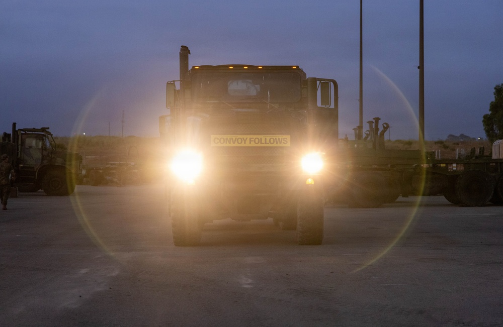 A military truck is photographed head-on at dusk or dawn with its headlights on, displaying a 'CONVOY FOLLOWS' sign on its front bumper. The bright headlights create a lens flare effect across the image. The vehicle is positioned in what appears to be a military staging or marshaling area with other military vehicles visible in the background.