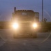 A military truck is photographed head-on at dusk or dawn with its headlights on, displaying a 'CONVOY FOLLOWS' sign on its front bumper. The bright headlights create a lens flare effect across the image. The vehicle is positioned in what appears to be a military staging or marshaling area with other military vehicles visible in the background.