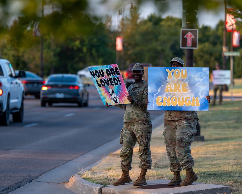 Two military personnel in camouflage uniforms stand on a roadside median holding encouraging signs reading 'You Are Loved!' and 'You Are Enough.' The scene appears to be a mental health awareness or morale-boosting initiative on what looks like a military installation road. Additional uniformed personnel with signs are visible in the background.