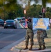 Two military personnel in camouflage uniforms stand on a roadside median holding encouraging signs reading 'You Are Loved!' and 'You Are Enough.' The scene appears to be a mental health awareness or morale-boosting initiative on what looks like a military installation road. Additional uniformed personnel with signs are visible in the background.
