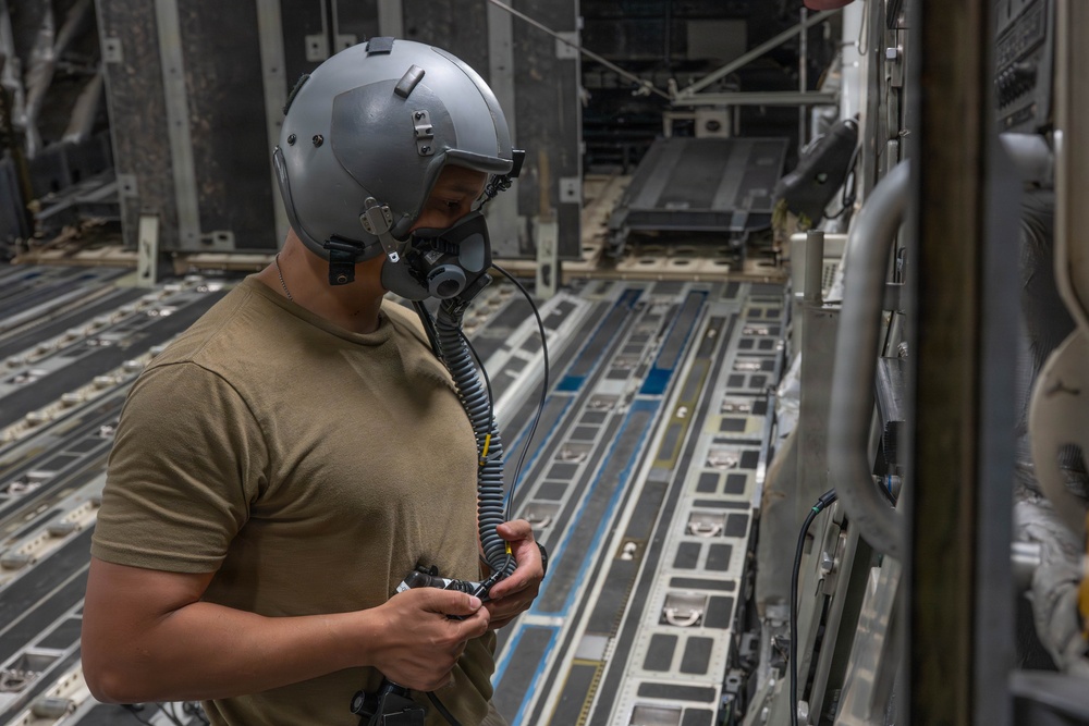 A military aircrew member wearing a flight helmet with oxygen mask and communications equipment stands inside the cargo hold of a large military transport aircraft. The individual appears to be inspecting or connecting communication cables while positioned along the interior sidewall of the aircraft. The cargo floor features roller conveyor tracks and tie-down rings typical of heavy-lift military transport aircraft.