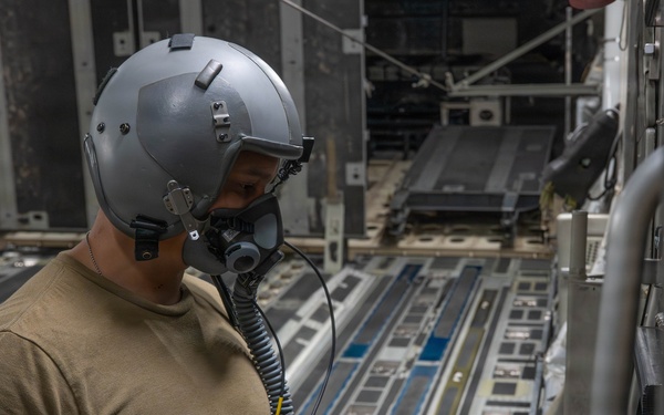 A military aircrew member wearing a flight helmet with oxygen mask and communications equipment stands inside the cargo hold of a large military transport aircraft. The individual appears to be inspecting or connecting communication cables while positioned along the interior sidewall of the aircraft. The cargo floor features roller conveyor tracks and tie-down rings typical of heavy-lift military transport aircraft.