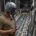 A military aircrew member wearing a flight helmet with oxygen mask and communications equipment stands inside the cargo hold of a large military transport aircraft. The individual appears to be inspecting or connecting communication cables while positioned along the interior sidewall of the aircraft. The cargo floor features roller conveyor tracks and tie-down rings typical of heavy-lift military transport aircraft.