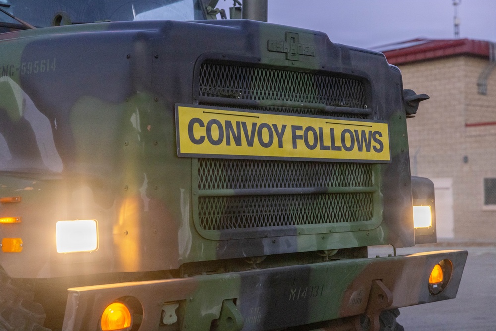 A military vehicle photographed from the front displays a prominent yellow sign reading 'CONVOY FOLLOWS,' indicating it is the lead or marker vehicle of a military convoy. The vehicle is painted in green camouflage and has its headlights and amber marker lights illuminated, suggesting operation in low-light conditions at dusk or dawn. A building with a red roof is partially visible in the background.
