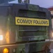 A military vehicle photographed from the front displays a prominent yellow sign reading 'CONVOY FOLLOWS,' indicating it is the lead or marker vehicle of a military convoy. The vehicle is painted in green camouflage and has its headlights and amber marker lights illuminated, suggesting operation in low-light conditions at dusk or dawn. A building with a red roof is partially visible in the background.