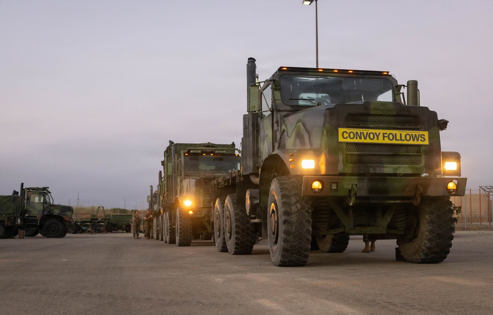 A military convoy of camouflage-painted heavy tactical trucks is staged on a paved surface at dusk or dawn. The lead vehicle prominently displays a yellow 'CONVOY FOLLOWS' sign on its front bumper, indicating it is the rear vehicle of a convoy formation. Multiple military personnel and additional military vehicles are visible in the background.