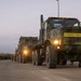 A military convoy of camouflage-painted heavy tactical trucks is staged on a paved surface at dusk or dawn. The lead vehicle prominently displays a yellow 'CONVOY FOLLOWS' sign on its front bumper, indicating it is the rear vehicle of a convoy formation. Multiple military personnel and additional military vehicles are visible in the background.