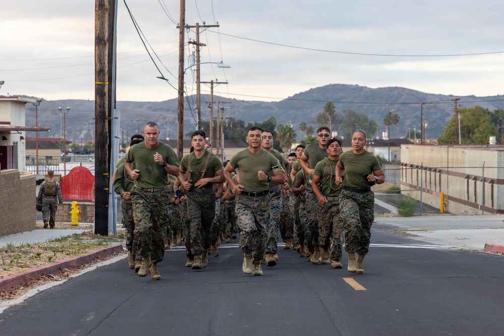 A group of U.S. Marines in uniform are conducting a formation run down a paved road on what appears to be a military base. The personnel are wearing standard MARPAT camouflage trousers and olive drab physical training shirts. The setting includes utility poles, fencing, low buildings, and arid hills in the background consistent with a Southern California military installation.