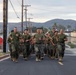 A group of U.S. Marines in uniform are conducting a formation run down a paved road on what appears to be a military base. The personnel are wearing standard MARPAT camouflage trousers and olive drab physical training shirts. The setting includes utility poles, fencing, low buildings, and arid hills in the background consistent with a Southern California military installation.