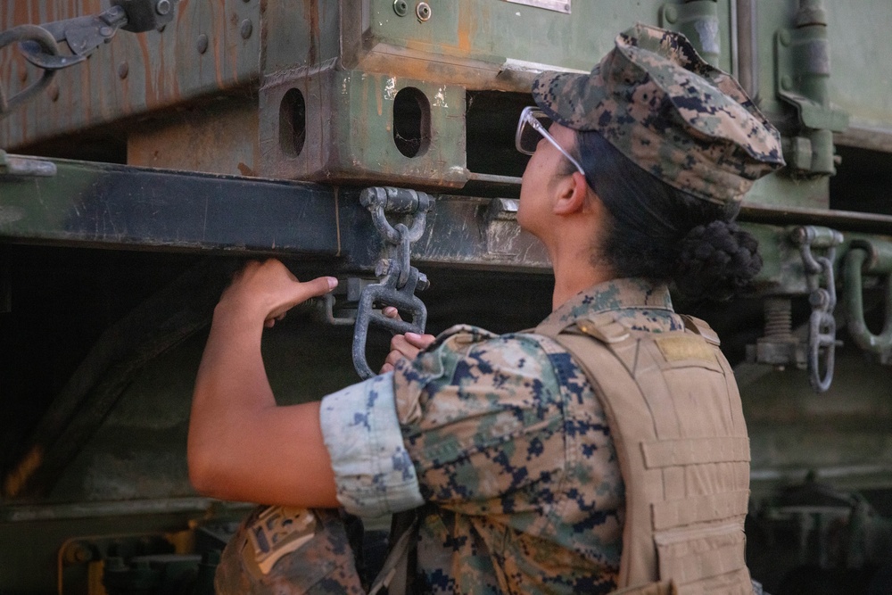 A female military service member in MARPAT (Marine Corps digital camouflage) uniform and tactical vest is inspecting or manipulating a shackle/clevis pin on a large military vehicle. She is wearing safety glasses and a patrol cap, and appears to be conducting a maintenance or rigging check on heavy equipment. The vehicle shows signs of wear with rust and weathering on its green painted surface.