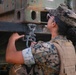 A female military service member in MARPAT (Marine Corps digital camouflage) uniform and tactical vest is inspecting or manipulating a shackle/clevis pin on a large military vehicle. She is wearing safety glasses and a patrol cap, and appears to be conducting a maintenance or rigging check on heavy equipment. The vehicle shows signs of wear with rust and weathering on its green painted surface.