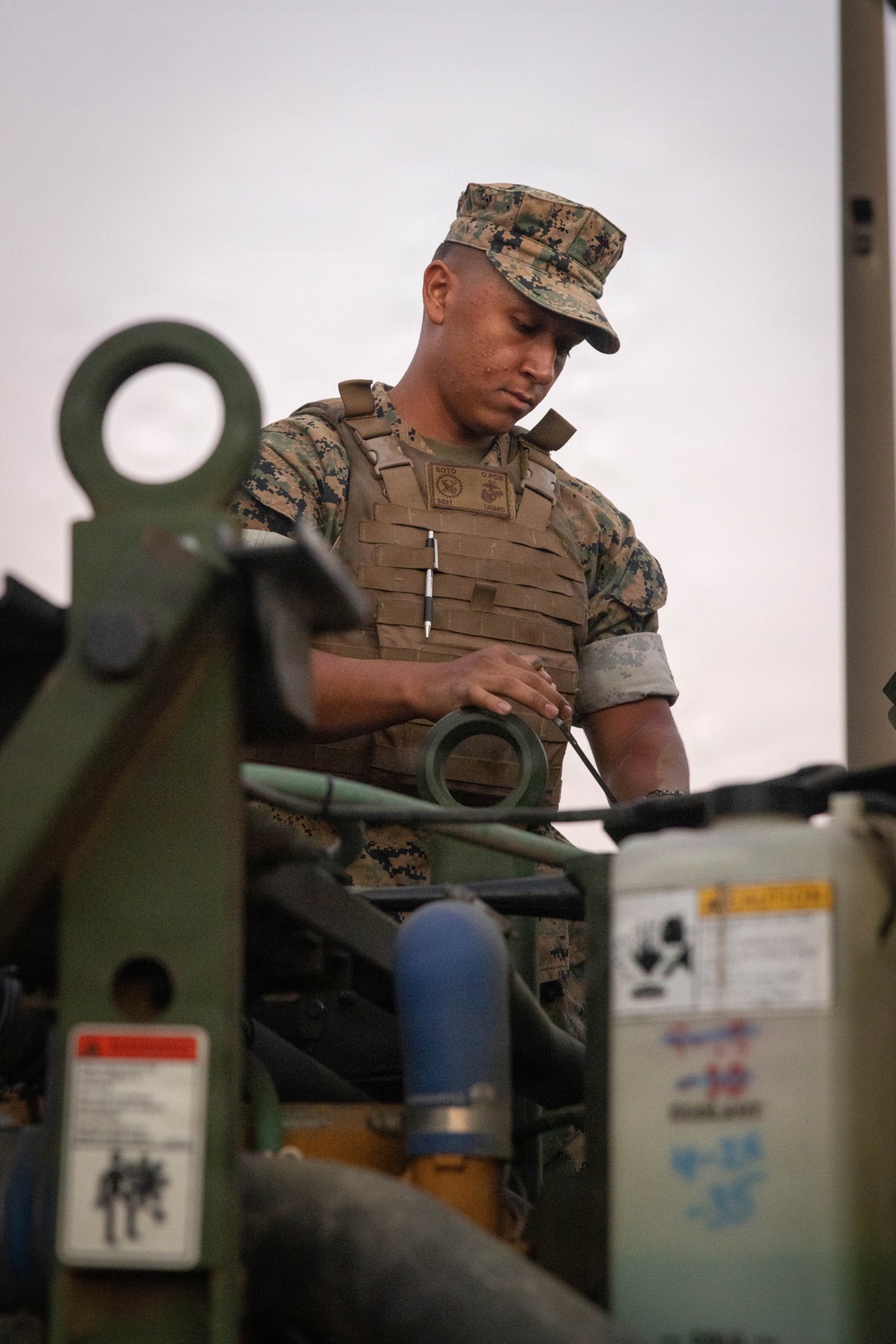 A U.S. Marine in MARPAT digital camouflage uniform and plate carrier vest is working with military equipment, appearing to inspect or operate a piece of machinery or vehicle. The Marine's name tape reads 'SOTO' and the USMC Eagle, Globe, and Anchor emblem is visible on the chest plate carrier. The image is taken at close range with military equipment in the foreground framing the subject.
