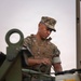 A U.S. Marine in MARPAT digital camouflage uniform and plate carrier vest is working with military equipment, appearing to inspect or operate a piece of machinery or vehicle. The Marine's name tape reads 'SOTO' and the USMC Eagle, Globe, and Anchor emblem is visible on the chest plate carrier. The image is taken at close range with military equipment in the foreground framing the subject.