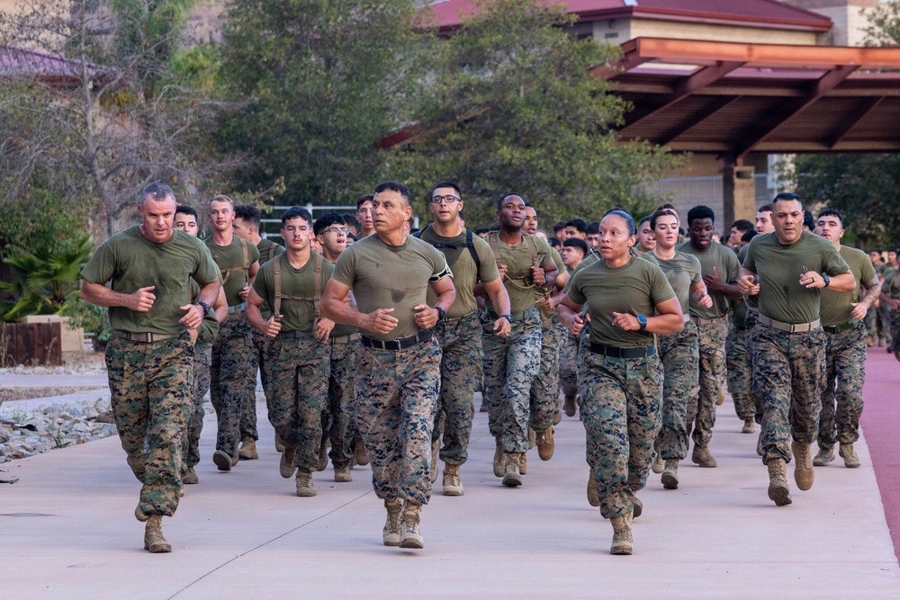 A large formation of military personnel in U.S. Marine Corps uniforms are conducting a group physical training run along a paved pathway. The personnel are wearing standard USMC MARPAT camouflage utilities with olive drab PT shirts and tan combat boots. Both male and female Marines are visible running in formation, suggesting a unit-level physical fitness event.