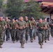 A large formation of military personnel in U.S. Marine Corps uniforms are conducting a group physical training run along a paved pathway. The personnel are wearing standard USMC MARPAT camouflage utilities with olive drab PT shirts and tan combat boots. Both male and female Marines are visible running in formation, suggesting a unit-level physical fitness event.