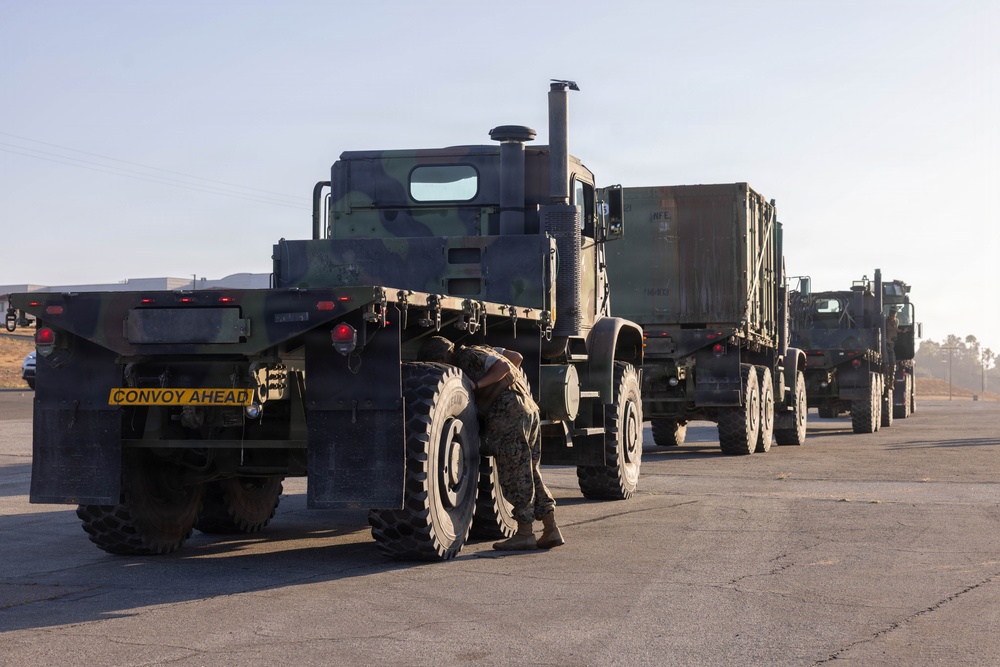 A military convoy of camouflage-painted heavy transport trucks is staged on a paved surface, likely a military base or airfield. A service member in uniform is crouched near the front wheel of the lead vehicle, appearing to perform an inspection or maintenance check. A yellow 'CONVOY AHEAD' warning sign is visible on the rear of the first flatbed trailer.
