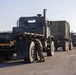 A military convoy of camouflage-painted heavy transport trucks is staged on a paved surface, likely a military base or airfield. A service member in uniform is crouched near the front wheel of the lead vehicle, appearing to perform an inspection or maintenance check. A yellow 'CONVOY AHEAD' warning sign is visible on the rear of the first flatbed trailer.