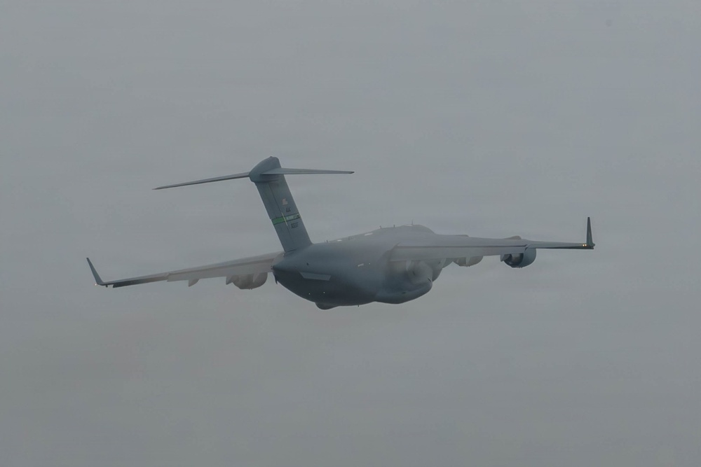 A large military cargo aircraft is photographed in flight against an overcast, grey sky. The aircraft displays a grey paint scheme consistent with military transport operations. The image was taken from a ground-level perspective looking upward at the aircraft as it passes overhead.