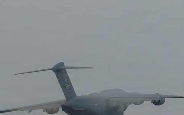 A large military cargo aircraft is photographed in flight against an overcast, grey sky. The aircraft displays a grey paint scheme consistent with military transport operations. The image was taken from a ground-level perspective looking upward at the aircraft as it passes overhead.