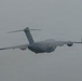 A large military cargo aircraft is photographed in flight against an overcast, grey sky. The aircraft displays a grey paint scheme consistent with military transport operations. The image was taken from a ground-level perspective looking upward at the aircraft as it passes overhead.