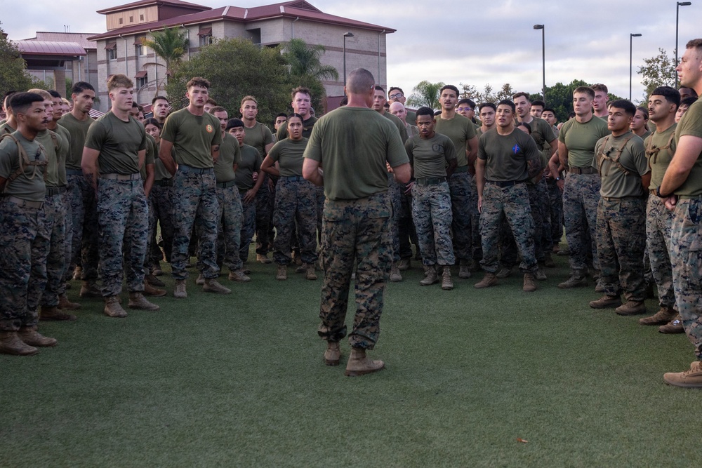 A military leader, likely a senior non-commissioned officer or officer, addresses a large formation of U.S. Marines on a grass field. The Marines are wearing standard MARPAT camouflage utilities and olive drab physical training shirts, gathered in a semicircle formation. The setting appears to be a military installation, with barracks-style buildings visible in the background.