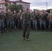 A military leader, likely a senior non-commissioned officer or officer, addresses a large formation of U.S. Marines on a grass field. The Marines are wearing standard MARPAT camouflage utilities and olive drab physical training shirts, gathered in a semicircle formation. The setting appears to be a military installation, with barracks-style buildings visible in the background.