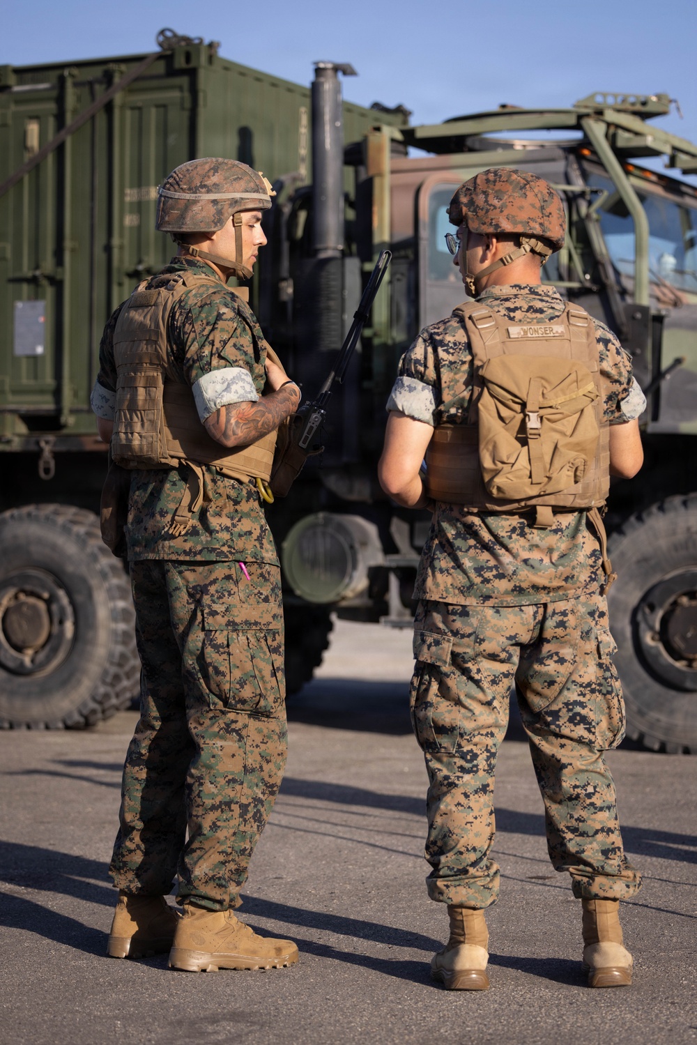 Two U.S. Marines in MARPAT (Marine Pattern) digital camouflage uniforms stand on a paved surface conversing in front of a military tactical truck. Both wear plate carrier vests, cover/helmets, and tan combat boots; one carries a rifle and wears a helmet while the other wears a patrol cap and has a name tape reading 'WONSER' visible on their vest. The setting appears to be a military installation or staging area under clear daylight conditions.