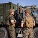 Two U.S. Marines in MARPAT (Marine Pattern) digital camouflage uniforms stand on a paved surface conversing in front of a military tactical truck. Both wear plate carrier vests, cover/helmets, and tan combat boots; one carries a rifle and wears a helmet while the other wears a patrol cap and has a name tape reading 'WONSER' visible on their vest. The setting appears to be a military installation or staging area under clear daylight conditions.