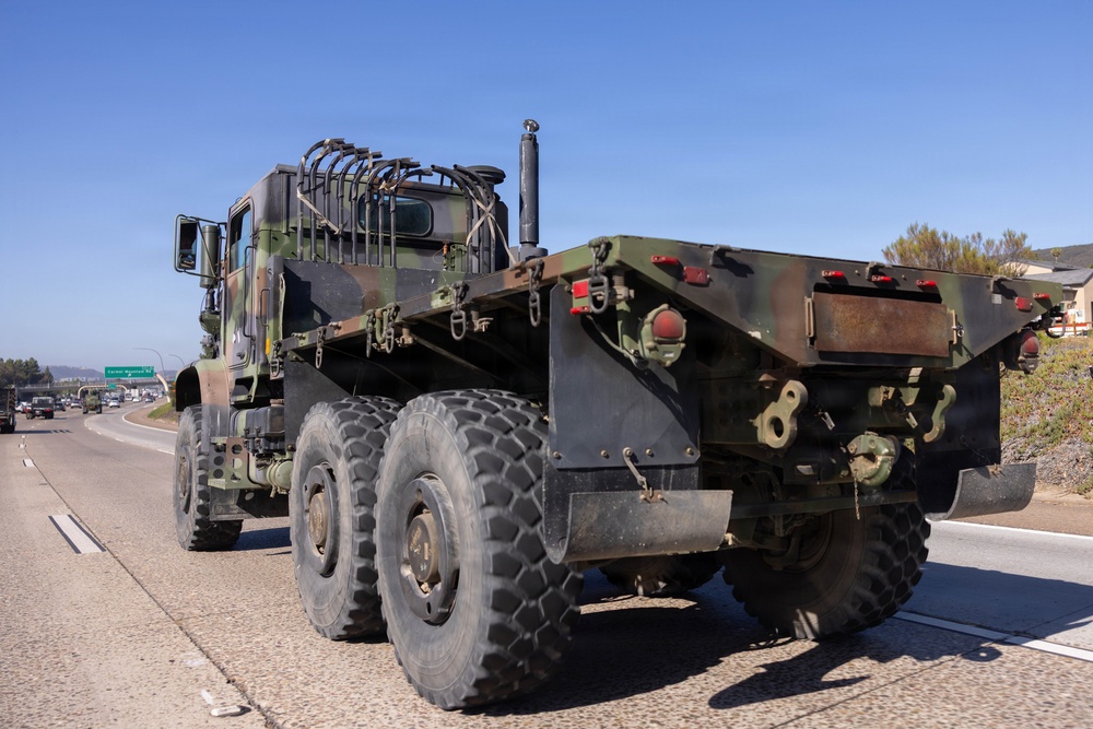 A military camouflage-painted heavy-duty truck, consistent with a US Army LMTV or HEMTT-type vehicle, is traveling on a multi-lane highway in what appears to be a sunny, clear-sky environment. The truck is viewed from the rear-left angle and appears to be a flatbed or cargo variant without a load. Additional vehicles and a green highway sign are visible in the background.