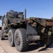A military camouflage-painted heavy-duty truck, consistent with a US Army LMTV or HEMTT-type vehicle, is traveling on a multi-lane highway in what appears to be a sunny, clear-sky environment. The truck is viewed from the rear-left angle and appears to be a flatbed or cargo variant without a load. Additional vehicles and a green highway sign are visible in the background.