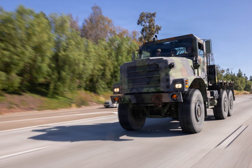 A large military truck in woodland camouflage paint scheme is traveling at speed on a multi-lane highway. The vehicle appears to be a heavy-duty tactical truck, likely a Medium Tactical Vehicle Replacement (MTVR) or similar platform, photographed with a panning technique that creates motion blur in the background. The setting appears to be a California highway based on the vegetation and road infrastructure.