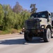 A large military truck in woodland camouflage paint scheme is traveling at speed on a multi-lane highway. The vehicle appears to be a heavy-duty tactical truck, likely a Medium Tactical Vehicle Replacement (MTVR) or similar platform, photographed with a panning technique that creates motion blur in the background. The setting appears to be a California highway based on the vegetation and road infrastructure.