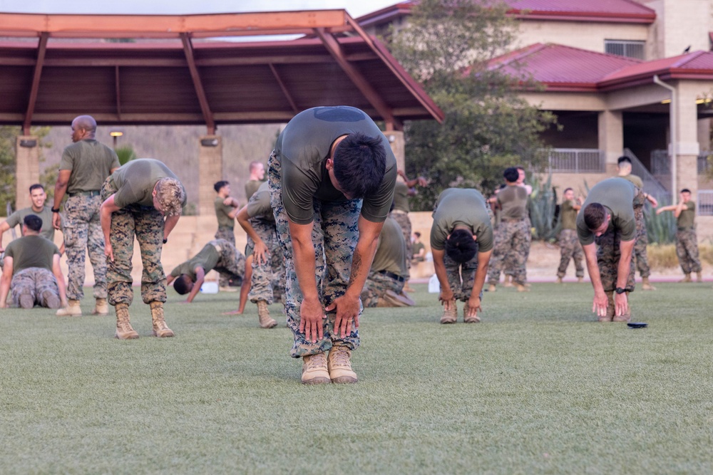 A large group of military personnel in U.S. Marine Corps uniforms are conducting a physical training session on a grass field. The soldiers are performing stretching and warm-up exercises, with the central figure bent forward touching his boots in a toe-touch stretch. The setting appears to be a military base facility with a covered pavilion structure visible in the background.