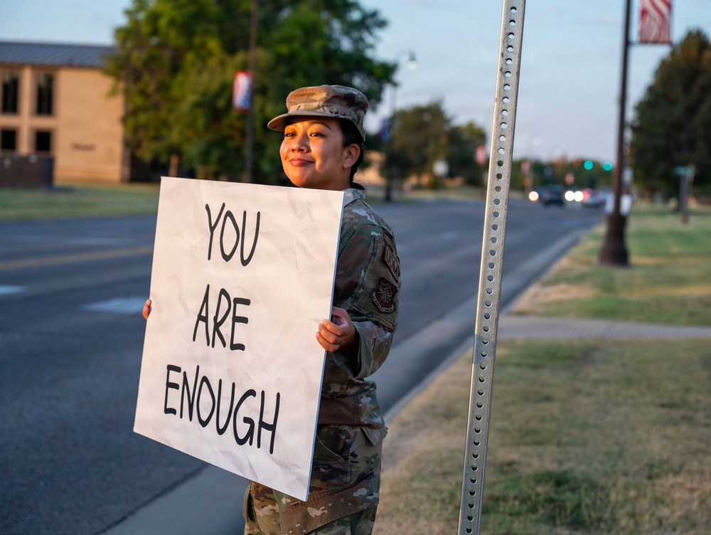 A person in military camouflage uniform and patrol cap stands along a roadside holding a large handwritten sign reading 'YOU ARE ENOUGH.' The setting appears to be a military installation at dusk or early morning, with an American flag visible in the background. The message on the sign is associated with mental health awareness and suicide prevention advocacy.