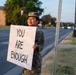 A person in military camouflage uniform and patrol cap stands along a roadside holding a large handwritten sign reading 'YOU ARE ENOUGH.' The setting appears to be a military installation at dusk or early morning, with an American flag visible in the background. The message on the sign is associated with mental health awareness and suicide prevention advocacy.